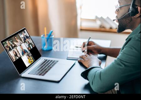 Virtuelle Konferenz mit multiethnischen Menschen. Afroamerikanischer Mann im Headset lernt aus der Ferne mit Laptop und Video-Chat-App. Auf dem Laptop-Bildschirm der Lehrer und Gruppe von verschiedenen Menschen, online Stockfoto