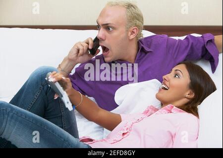 Ein junger Mann, der ein Mobiltelefon auf dem Bett benutzte und eine junge Frau sich auf ihn lehnte Stockfoto