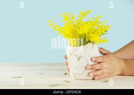 Muttertagsgeschenk. Kinderhände halten eine diy Keramik Vase mit gelben Blumen. Isoliert auf blauem Hintergrund. Stockfoto