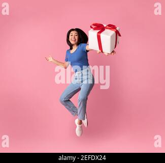 Volle Länge vertikale Foto von springen hoch aufgeregt asiatische Dame halten und zeigen Geschenk-Box in der Nähe der Kamera, Geschenk isoliert über pastellrosa s Stockfoto