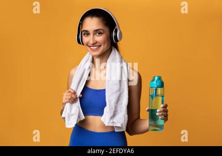 Wellness- Und Trainingskonzept. Porträt einer fröhlichen, sportlichen jungen Frau, die nach dem Training im Fitnessstudio eine Flasche mit Mineralwasser hält und Köpfe trägt Stockfoto