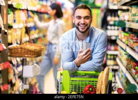 Porträt eines lächelnden jungen Casual-Mannes, der sich im Innenbereich auf dem Einkaufswagen des Einkaufswagens stützt, sein Smartphone benutzt, posiert und die Kamera anschaut. Zufriedener männlicher Kunde Stockfoto