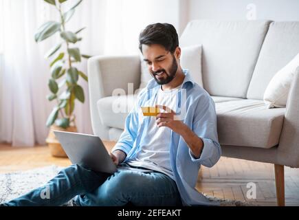 Millennial Arab Guy Macht Online-Zahlungen Mit Laptop Und Kreditkarte Zu Hause, Sitzen Auf Dem Boden In Gemütlichen Wohnzimmer, Young Eastern Man Einkaufen In Stockfoto