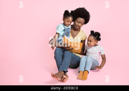 Glückliche Familie, junge afrikanische Frau Mutter, Buch mit faszinierenden Märchen zu ihren beiden niedlichen kleinen Töchtern sitzen auf dem Boden auf isoliert Stockfoto