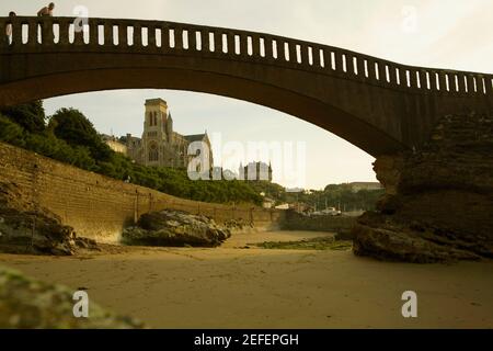 Bogenbrücke am Strand und eine Kathedrale im Hintergrund, Port des Pecheurs, Eglise Sainte Eugenie, Biarritz, Frankreich Stockfoto