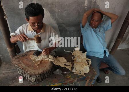 Handwerker macht eine Lederpuppe für wayang kulis Theater im klassischen Wayang Handwerksladen in Yogyakarta, Zentral Java, Indonesien. Rechts ist der Bastelmanager Dodo mit einem blauen Hemd abgebildet. Das traditionelle Puppenschattentheater Wayang kulit ist auf den Inseln Java und Bali in Indonesien weit verbreitet. Puppen für Wayang kulis sind aus Büffelleder gefertigt. Die Herstellung einer Puppe dauert etwa sechs Tage. Jeder Charakter hat seine eigenen Eigenschaften und kann leicht von den Zuschauern identifiziert werden. Stockfoto