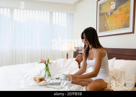 Junge Frau, die mit einem mittleren Erwachsenen im Bett frühstückte Mann, der neben ihr schläft Stockfoto