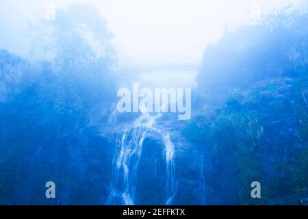 Mystische Holzbrücke über einen Wasserfall in blauem Nebel. Sapa, Vietnam, Blick von unten auf die Brücke. Stockfoto