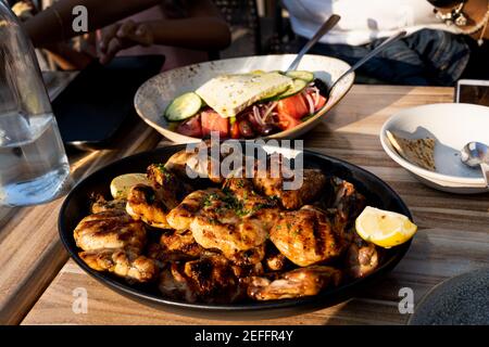 Authentisches Foto von griechischem gegrilltem Huhn mit außer Fokus griechischem Salat im Hintergrund unter goldenem Licht Sonnenlicht. Menschen können gesehen werden Stockfoto