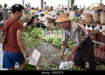 YANGON, MYANMAR - DEZEMBER 31 2019: Zwei burmesische Männer tauschen Geld aus, wenn sie Gemüse und Obst auf einem Straßenmarkt kaufen und verkaufen Stockfoto