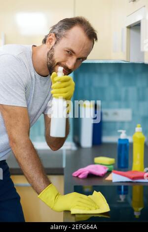 Porträt von professionellen männlichen Reiniger tragen Handschuhe Blick auf Kamera Und mit Sprühwaschmittel während der Reinigung der Oberfläche in der Küche Stockfoto
