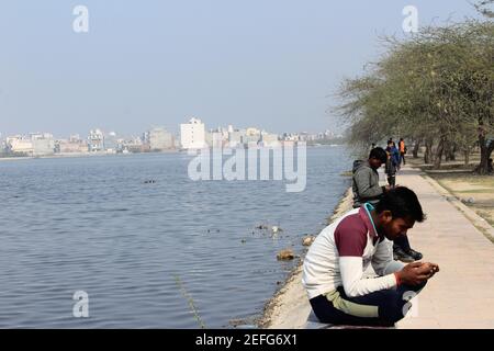 Kleiner indischer Junge, der sein Handy anschaut, während er an einem sonnigen Winternachmittag in der Nähe des Bhalswaer Horseshoe Sees in Delhi, Indien sitzt Stockfoto