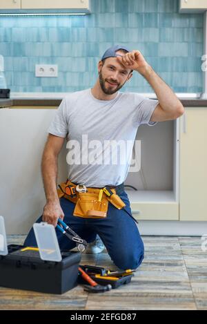 Qualitativ hochwertige Arbeit. Attraktive junge Handwerker, Klempner trägt Werkzeug Gürtel lächelnd an der Kamera, während sie einen Rohrschlüssel, bereit für die Befestigung Waschbecken Rohr in der Stockfoto