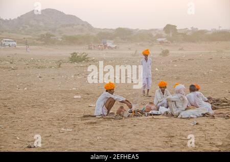 Während der Pushkar Messe sitzen eine Gruppe von Kamelhandlern auf dem Messegelände in der Abendsonne. Der orangefarbene Turban auf ihren Köpfen ist hell. Stockfoto