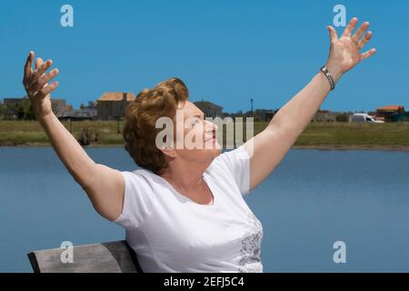 Ältere Frau sitzt am See mit ausgestreckten Armen Stockfoto