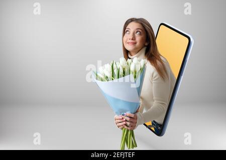 Lächelnd schönes Mädchen schaut aus einem Smartphone und hält einen großen Strauß von weißen Tulpen. Junge Frau online auf einem Handy-Bildschirm. Speicherplatz kopieren. Stockfoto