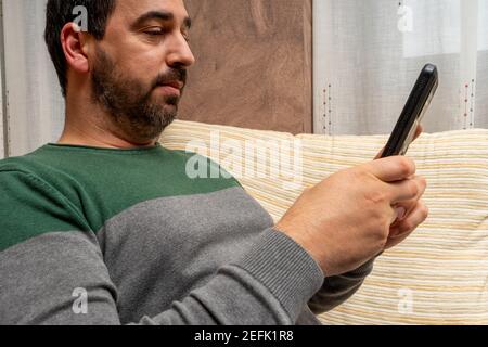 Bärtiger Mann in einem grauen und grünen Pullover sitzt auf der Couch zu Hause mit seinem Smartphone. Technologiekonzept Stockfoto