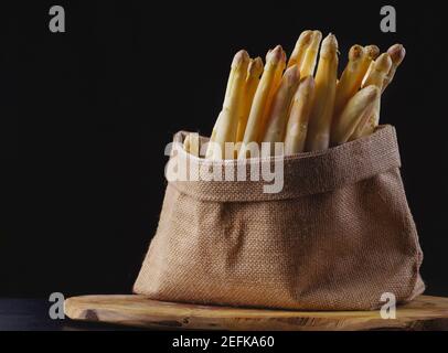 Frisch gepflückte rohe weiße Spargelbündel aus biologischem Anbau.Weißer Spargel auf schwarzem Hintergrund in einem Stoffbeutel auf einem hölzernen Küchbrett, Gemüse, gesund Stockfoto