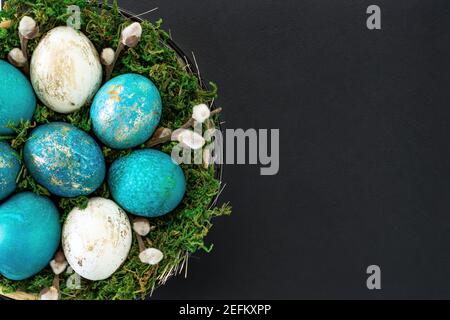 Osterhintergrund mit einem Nest mit farbigen Eiern Nahaufnahme auf einem schwarzen Hintergrund mit Kopierraum. Türkis und Gold Eier liegen in einem Korb mit Moos und Stockfoto