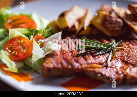 Gegrilltes Schweinefleisch mit gebackenen Kartoffeln und mit Salat. Das Essen wird auf einem Teller serviert. Köstliches gegrilltes Fleisch. Gebratenes Schweinesteak auf dem weißen Teller. Stockfoto