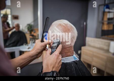 Barbier schneidet einem alten Kunden, einem herrn mit weißen Haaren, in seinem Friseurhaus Haare Stockfoto