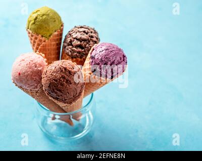 Verschiedene Eisschaufeln in Zapfen mit Kopierraum. Bunte Eis in Zapfen Schokolade, Erdbeere, Heidelbeere, Pistazie oder Matcha, Kekse Schokolade Sandwich-Kekse auf blauem Hintergrund Stockfoto