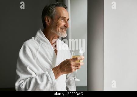 Lächelnder älterer Mann mit Weinglas. Seitenansicht eines bärtigen, reifen Mannes, der einen Bademantel in einem Hotelzimmer trägt Stockfoto