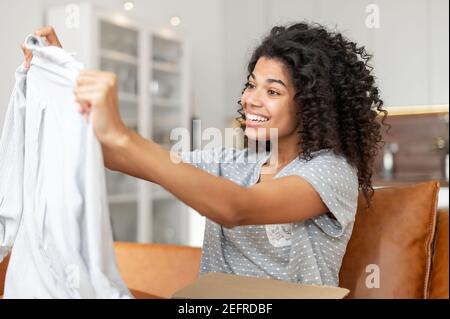 Zufriedene Biracial junge Frau mit einer Afro Frisur Auspacken Paket-Box sitzt auf dem Sofa zu Hause, aufgeregt afrikanische Frau schaut auf gelieferte Ware Stockfoto