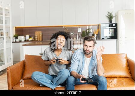 Ein multirassisches Paar mit Joysticks sitzt auf der Couch zu Hause und spielt Videospiel, eine afrikanische Frau ist Gewinner, schreien glücklich, während ihr Freund Stockfoto
