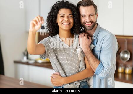 Fröhliche junge multi ethnischen Paar in der Liebe stehen in umarmt die Küche, hält die Schlüssel eines neuen Hauses. Freudige internationale Neuvermählte sind neu eingezogen Stockfoto