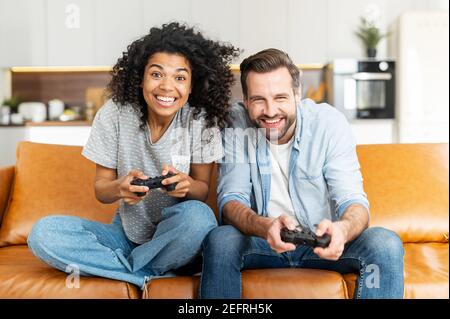 Ein multirassisches Paar genießt Zeit spielen Videospiel auf der Konsole, aufgeregt und angespannt junge afrikanische Frau und kaukasischen Kerl mit einem Joysticks versuchen, Stockfoto