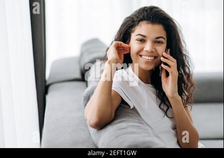 Lächelnd multirassische schwarze lockige Mädchen nutzt Handy, im Gespräch mit Freunden oder Familie, während sitzt auf der Couch im Wohnzimmer und schaut auf Kamera, Kommunikationskonzept Stockfoto