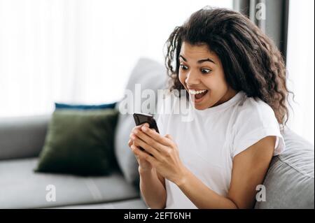 Glückliche Freiberuflerin ist überglücklich. Fröhliche junge afroamerikanische Frau sitzt auf dem Sofa, benutzt Smartphone, bekommt gute Nachrichten, schaut freudig auf den Telefonbildschirm Stockfoto