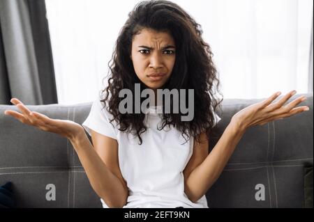 Lächelnd multirassische schwarze lockige Mädchen nutzt Handy, im Gespräch mit Freunden oder Familie, während sitzt auf der Couch im Wohnzimmer und schaut auf Kamera, Kommunikationskonzept Stockfoto