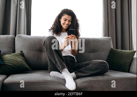 Moderne stilvolle Mädchen Bloggen in sozialen Medien, mit Smartphone. Hübsche afroamerikanische Frau, die mit Freunden oder Familie chattet und lächelt, während sie auf der Couch im Wohnzimmer sitzt und im Internet surft Stockfoto