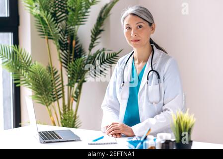 Grauhaarige weibliche Allgemeinärztin mittleren Alters, in medizinischer Uniform gekleidet, schaut direkt auf die Kamera am Arbeitsplatz in der Klinik. Ärztliche Beratung, Gesundheitskonzept Stockfoto