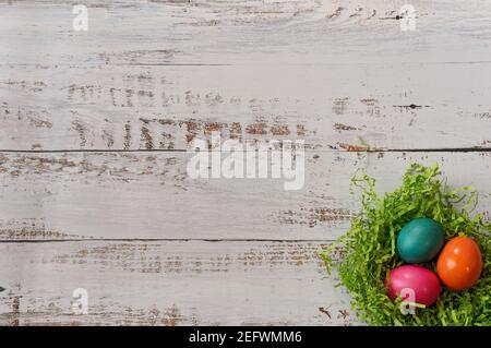 Drei bunt bemalte ostereier in einem ostergrasnest in der unteren rechten Ecke eines weißen Holzhintergrunds, mit Kopierraum Stockfoto