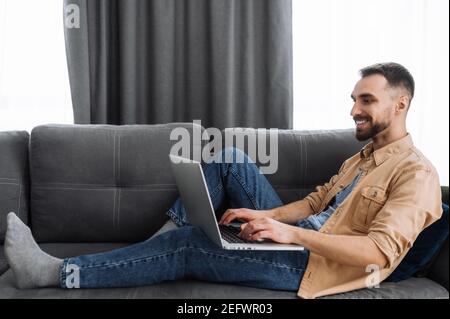 Kaukasischer männlicher Freiberufler sitzt auf dem Sofa und chillt zu Hause. Bärtiger Kerl in Freizeitkleidung verwenden Sie einen Laptop, Filme oder lustige Videos, oder arbeiten von zu Hause aus und lächeln Stockfoto
