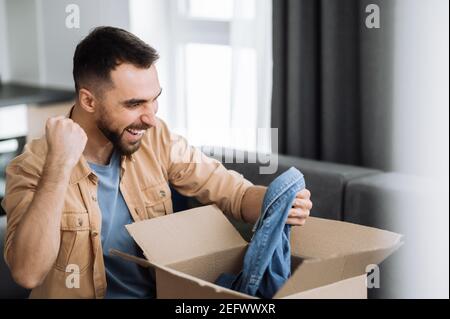 Überglücklich glücklicher Mann packte sein Paket aus, glücklich darüber, eine lange erwartete Bestellung zu bekommen. Kaukasischen modernen Kerl Einkaufen in Internet-Shops, Kauf neuer Kleidung online, Online-Shopping-Konzept Stockfoto
