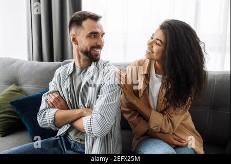 Fröhliches Ehepaar, das zu Hause chillt und auf dem Sofa sitzt. Kaukasischen Mann schaut mit Liebe auf ihre schöne afroamerikanische Frau, Wochenende zusammen Stockfoto