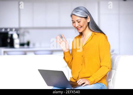 Lächelnd freundliche asiatische grauhaarige ältere Dame, sitzt auf der Couch im Wohnzimmer, mit Laptop, während der Arbeit aus der Ferne, kommuniziert mit Geschäftskollegen oder Freunden, Wellen Hand, Grüße Stockfoto