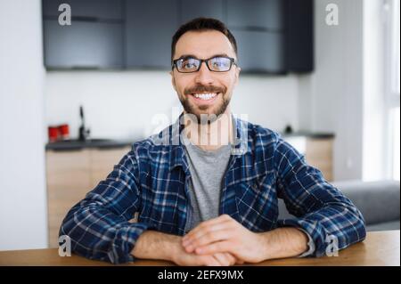 Webcam-Porträt von glücklich Freiberufler Kerl während der Videokonferenz, er schaut auf die Kamera und lächelt. Junge Erwachsene stilvolle intelligente Mann trägt Brillen entfernt arbeitet von zu Hause Stockfoto