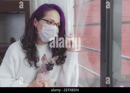 Eine unter Quarantäne gestellte Frau schaut durch das Fenster, sie hat ihre Gesichtsmaske aus der globalen Pandemie Stockfoto