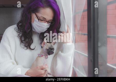 Eine unter Quarantäne gestellte Frau schaut durch das Fenster, sie hat ihre Gesichtsmaske aus der globalen Pandemie Stockfoto