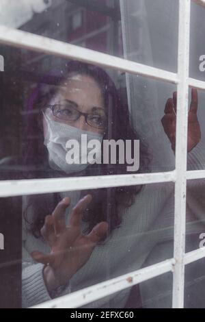 Eine unter Quarantäne gestellte Frau schaut durch das Fenster, sie hat ihre Gesichtsmaske aus der globalen Pandemie Stockfoto