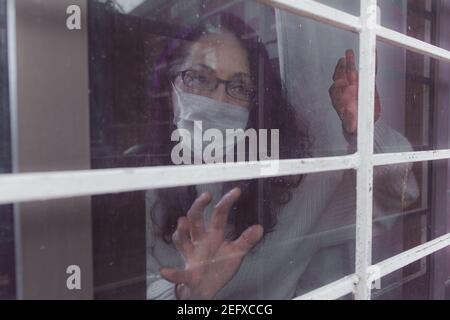 Eine unter Quarantäne gestellte Frau schaut durch das Fenster, sie hat ihre Gesichtsmaske aus der globalen Pandemie Stockfoto