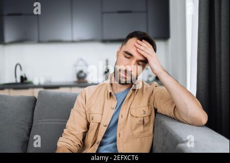 Müde kaukasischer bärtiger Mann sitzt auf dem Sofa und hat Kopfschmerzen. Ungesunder Kerl fangen eine Erkältung oder mit Grippe, verärgert über Krankheit, unglücklicher Mann fühlen sich depressiv Stockfoto