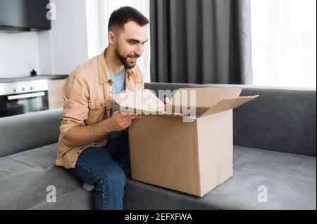 Lieferung nach Hause. Stilvoller Kaukasianer bekam seine Bestellung, packte ein Paket aus. Glücklicher Mann mit Interesse Blick in Karton, sitzen auf dem Sofa, der Kerl Einkaufen in Internet-Shops, Kauf neuer Kleidung online Stockfoto