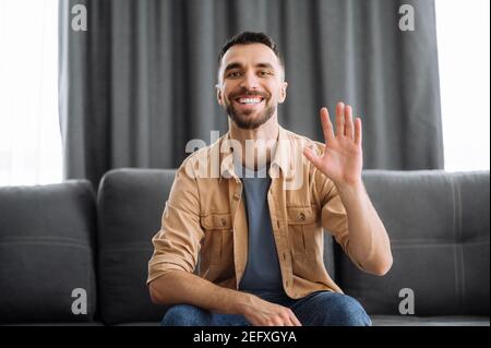 Porträt eines fröhlichen kaukasischen Freiberuflers während der Videokonferenz, er schaut auf die Kamera winkend Hand und lächelnd, Gruß seiner Freunde, Familie oder Kollegen, sitzen auf einer Couch Stockfoto