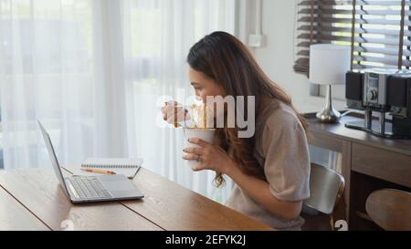 Asiatische Geschäftsfrau essen Instant Nudeln während der Arbeit auf Laptop-Computer zu Hause Büro, glücklich schöne junge Frau sitzen auf Schreibtisch Arbeit overtim Stockfoto